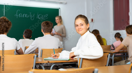 Portrait of positive teenage schoolgirl sitting on lesson in classroom