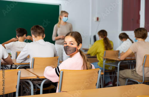 Young girl in face mask turned around and looking at camera while sitting at desk during lesson in classroom.