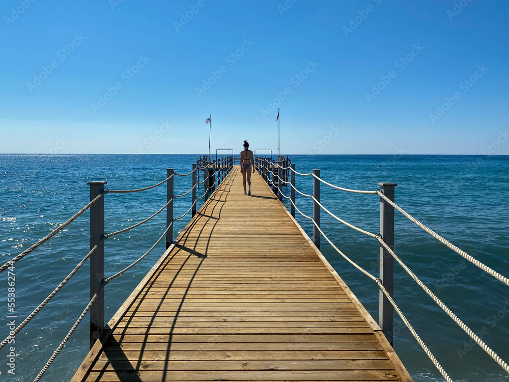 Naklejka premium A girl on a wooden long pontoon, a pier with rope railings on the sea on the beach on vacation in a heavenly warm eastern tropical country resort