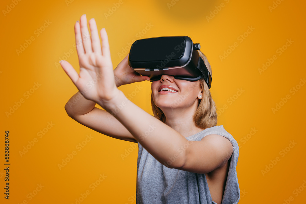 Attractive woman using virtual reality glasses, stretching out her palm, trying to touch something, isolated on yellow background. VR headset. Future