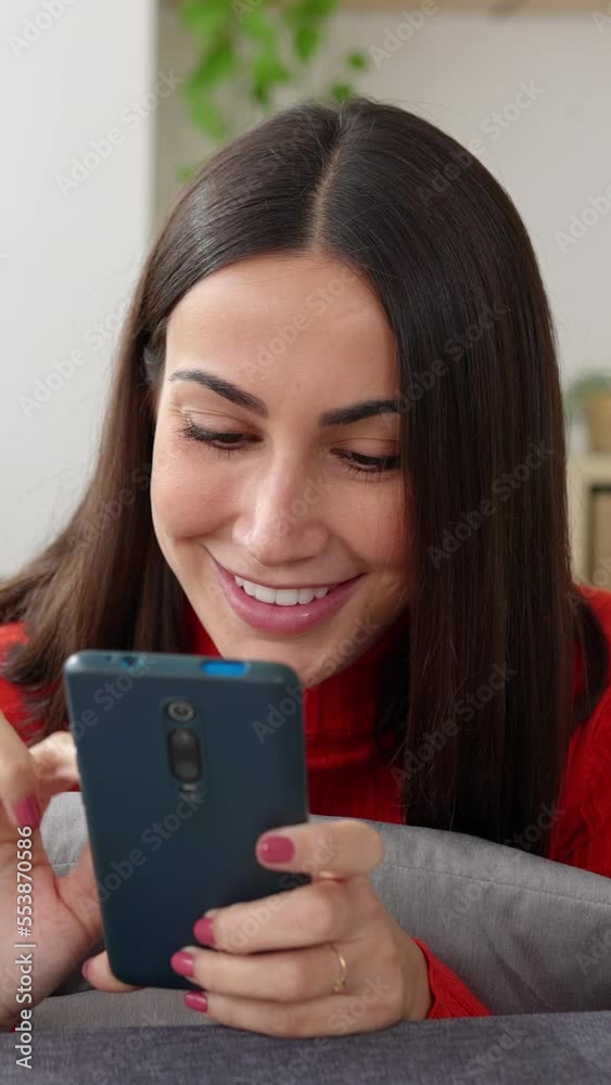 Vertical video shot of cheerful young pretty woman using mobile phone while relaxing on sofa at home. Smiling attractive female lying on couch typing message on smartphone. Vertical video shot of cheerful young pretty woman using mobile phone while relaxing on sofa at home. Smiling attractive female lying on couch typing message on smartphone.