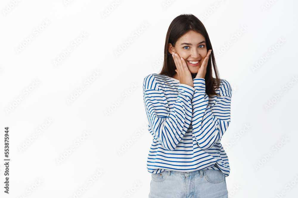 Image of smiling beautiful girl touched her healthy clear skin and looks pleased with cosmetic skincare product result, stands over white background Image of smiling beautiful girl touched her healthy clear skin and looks pleased with cosmetic skincare product result, stands over white background