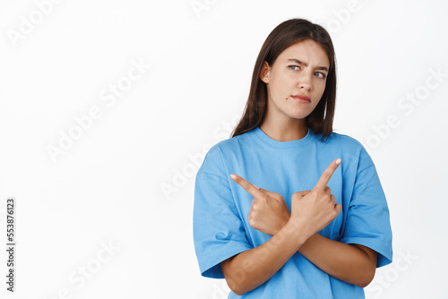 Thoughtful young modern girl in t-shirt makes choice, points sideways and looking with pondering serious face expression left, standing over white background © Cookie Studio