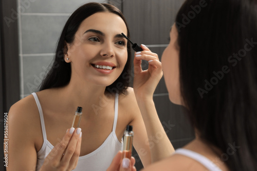Young woman applying oil onto eyelashes near mirror indoors