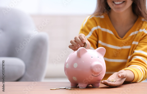 Woman putting coin into piggy bank at wooden table, closeup. Space for text