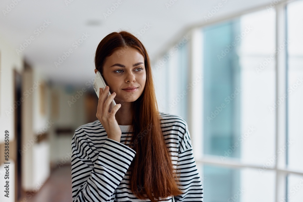 Fototapeta premium A woman talking on the phone in the office of a building and looking out the window at the big city
