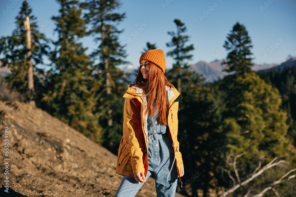 Woman hiking in the mountains in the autumn, a smile and happiness in a yellow cape with red hair full-length standing in front of trees and mountains in the sunset light