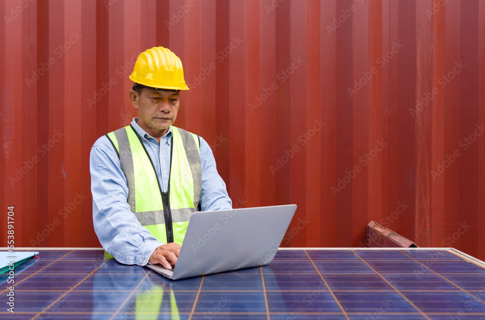 Senior contractor in safety vest and yellow hardhat typing on laptop computer keyboard. Check out the delivery schedule planning for the next shipment. He use solar cell panel as a table. Senior contractor in safety vest and yellow hardhat typing on laptop computer keyboard. Check out the delivery schedule planning for the next shipment. He use solar cell panel as a table.