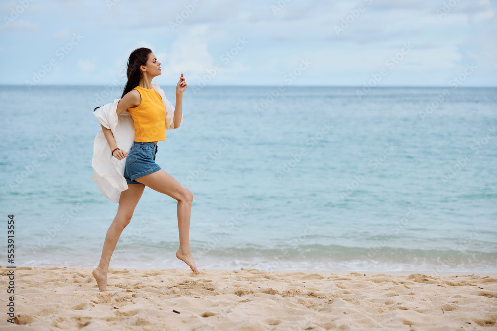 Sports woman runs along the beach in summer clothes on the sand in a yellow tank top and denim shorts white shirt flying hair ocean view, beach vacation and travel Sports woman runs along the beach in summer clothes on the sand in a yellow tank top and denim shorts white shirt flying hair ocean view, beach vacation and travel
