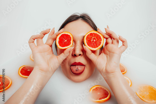 A girl in a bath filled with milk closes her eyes with orange slices and shows her tongue
