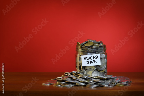 A glass Swear Jar overflowing with Australian coins to illustrate the excessive use of bad language, right of frame on a vibrant red background; captured in a studio
