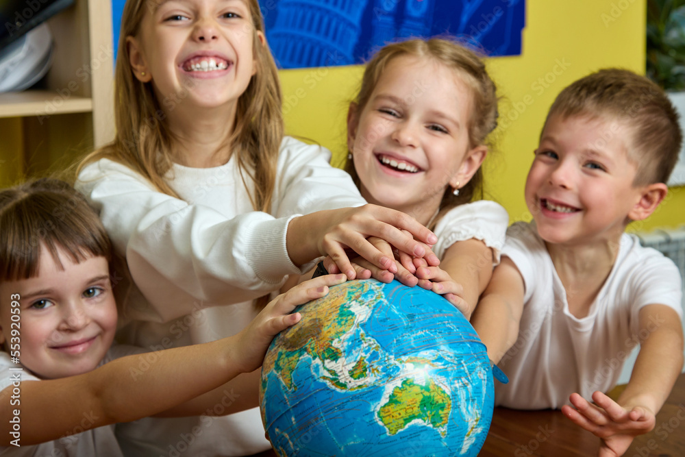 Happy children put their hands together on globe in school's office. Against background of yellow wall, cheerful schoolchildren folded their hands on globe standing on table as a sign of friendship Happy children put their hands together on globe in school's office. Against background of yellow wall, cheerful schoolchildren folded their hands on globe standing on table as a sign of friendship