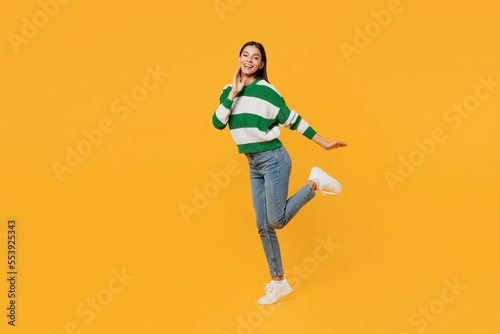 Full body side view smiling fun young latin woman in casual cozy green knitted sweater look camera raise up leg hold face isolated on plain yellow background studio portrait People lifestyle concept
