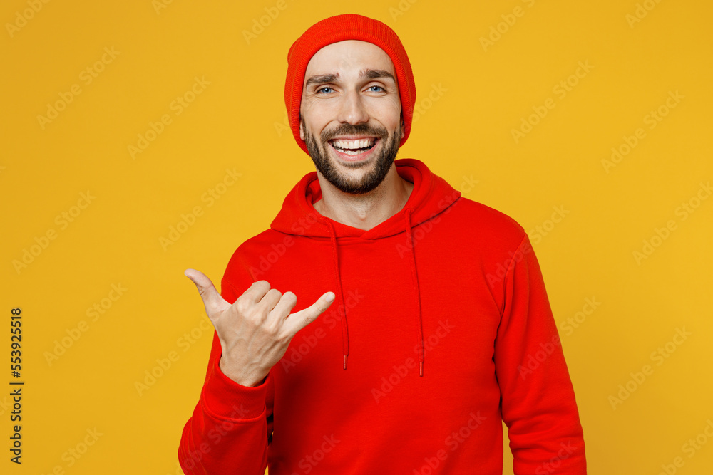 Young smiling cheerful fun happy cool optimist caucasian man wear red hoody hat look camera showing shaka gesture isolated on plain yellow color background studio portrait. People lifestyle concept. Young smiling cheerful fun happy cool optimist caucasian man wear red hoody hat look camera showing shaka gesture isolated on plain yellow color background studio portrait. People lifestyle concept.