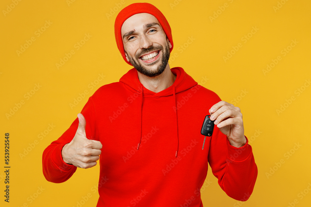 Young overjoyed man wear red hoody hat look camera hold car key fob keyless system do winner gesture show thumb up isolated on plain yellow color background studio portrait. People lifestyle concept. Young overjoyed man wear red hoody hat look camera hold car key fob keyless system do winner gesture show thumb up isolated on plain yellow color background studio portrait. People lifestyle concept.