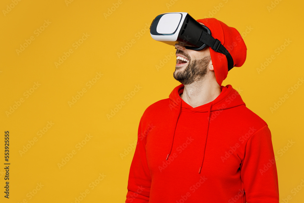 Side view young amazed smiling fun impressed caucasian man wearing red hoody hat watching in vr headset pc gadget isolated on plain yellow color background studio portrait. People lifestyle concept.