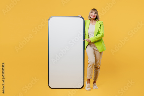 Full body elderly woman 50s years old wear green jacket white t-shirt stand near big huge blank screen mobile cell phone show smartphone with area isolated on plain yellow background studio portrait.