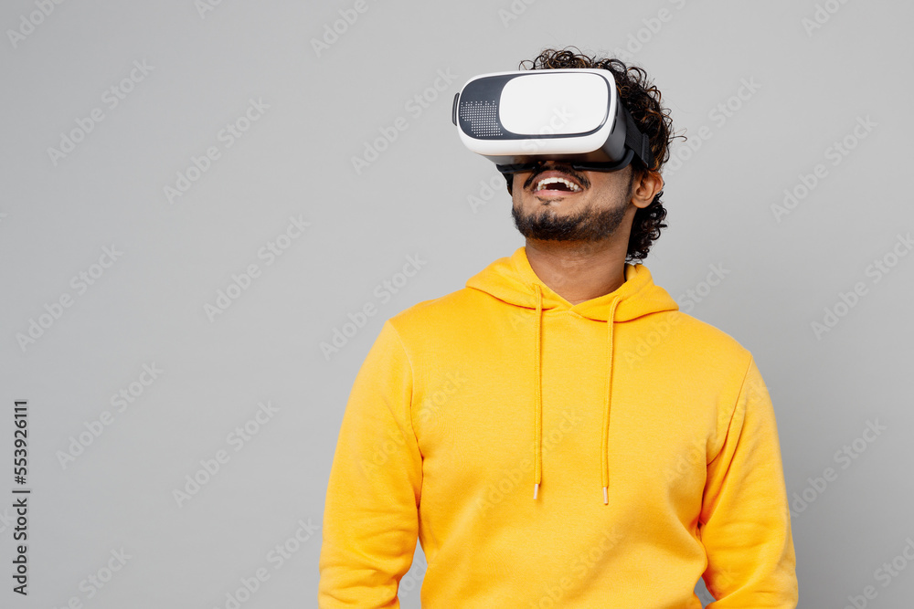 Young gambling fun cool happy smiling Indian man 20s he wearing casual yellow hoody watching in vr headset pc gadget isolated on plain grey color background studio portrait. People lifestyle portrait.