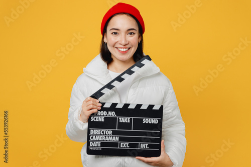 Young cheerful woman of Asian ethnicity wear white padded windbreaker jacket red hat hold in hand classic black film making clapperboard isolated on plain yellow background. People lifestyle concept.