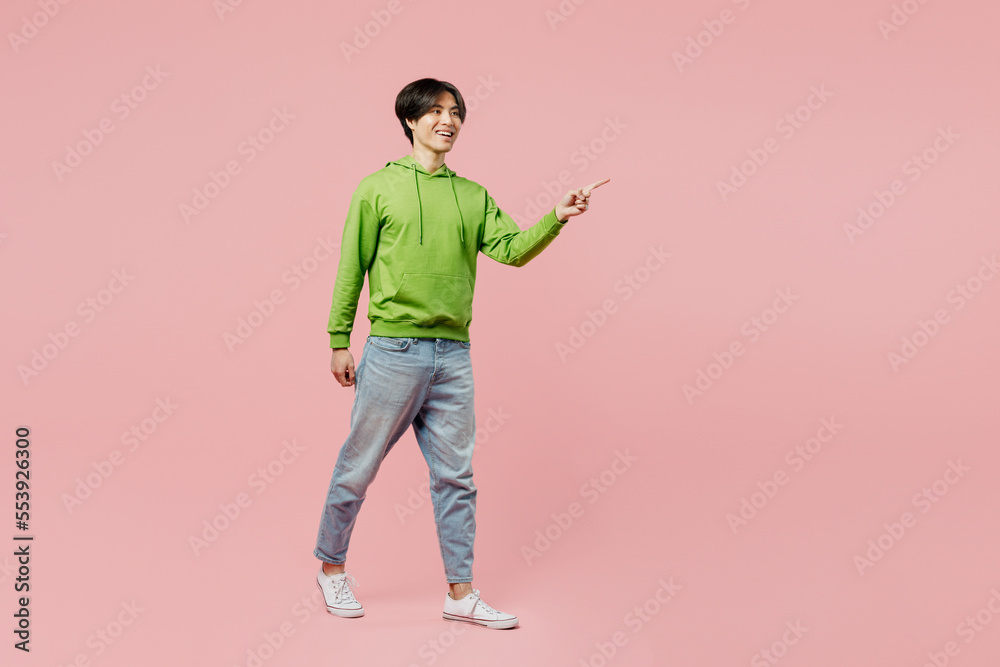 Full body sideways fun young man of Asian ethnicity wear green hoody walking going strolling point index finger aside on area workspace isolated on plain pastel light pink background studio portrait. Full body sideways fun young man of Asian ethnicity wear green hoody walking going strolling point index finger aside on area workspace isolated on plain pastel light pink background studio portrait.