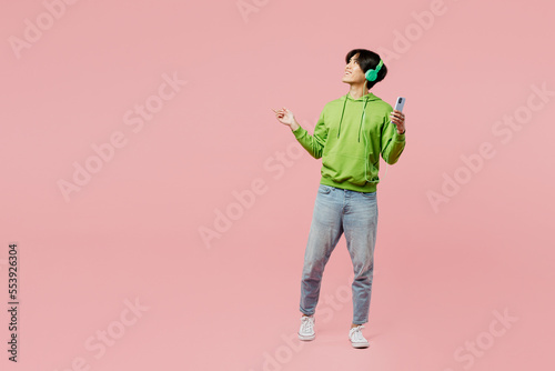 Full body young man of Asian ethnicity wear green hoody headphones listen to music use mobile cell phone point aside on area isolated on plain pastel light pink background. People lifestyle concept.