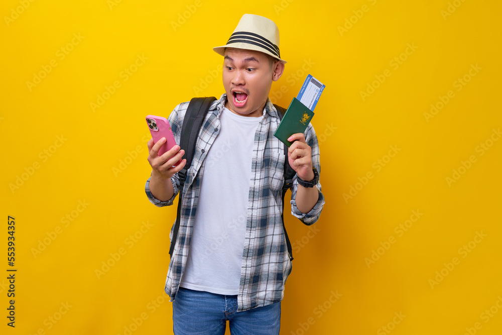 Excited young traveler tourist Asian man wearing casual clothes hat with backpack holding passport ticket mobile cell phone isolated on yellow background. Air flight journey concept