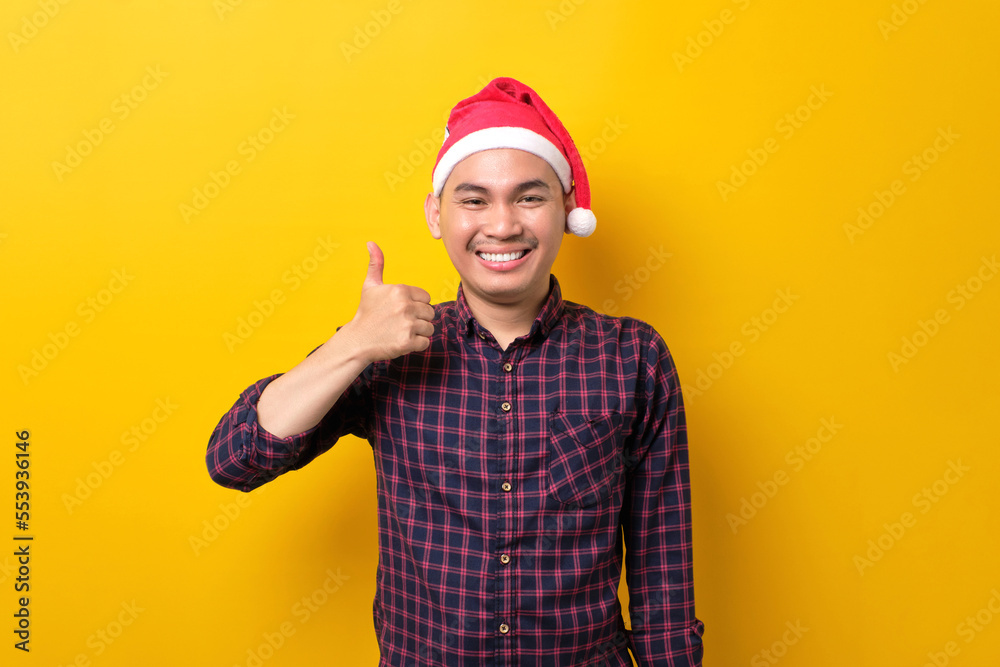 Cheerful young Asian man in Santa hat showing thumbs up, looking at camera on yellow studio background. Happy New Year 2023 celebration merry holiday concept