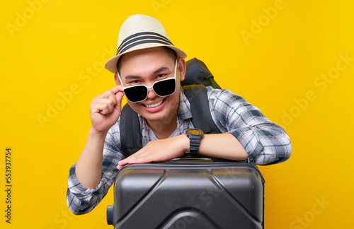 Smiling traveler tourist Asian man in casual clothes hat backpack glasses leaning on suitcase valise isolated on yellow background. Passenger travel abroad weekends getaway Air flight journey concept © Bangun Stock Photo