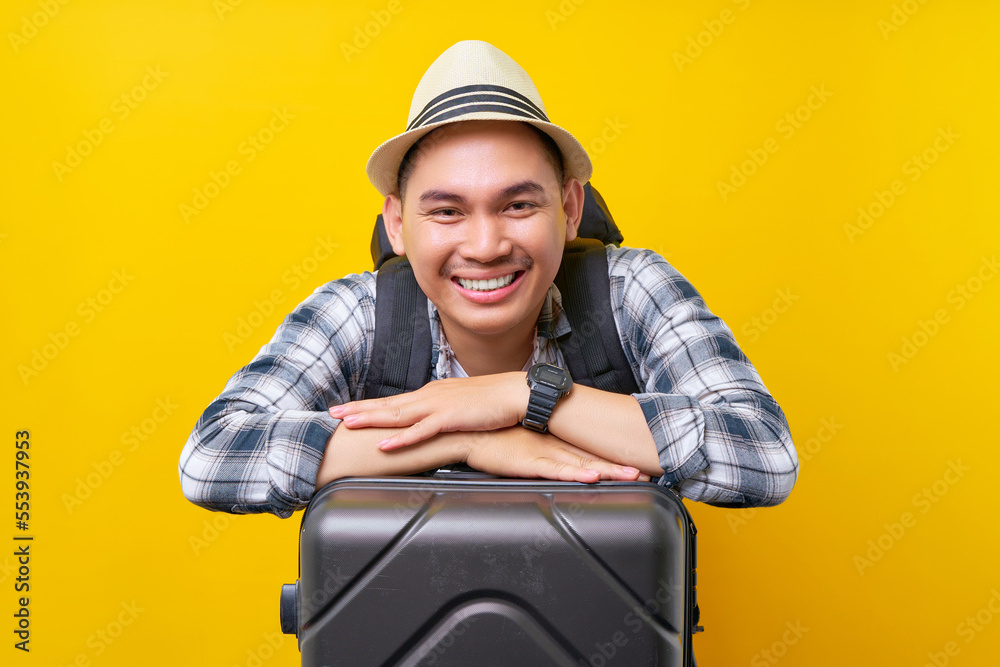 Smiling traveler tourist Asian man in casual clothes hat and backpack leaning on grey suitcase valise on yellow background. Passenger travel abroad weekends getaway Air flight journey concept Smiling traveler tourist Asian man in casual clothes hat and backpack leaning on grey suitcase valise on yellow background. Passenger travel abroad weekends getaway Air flight journey concept