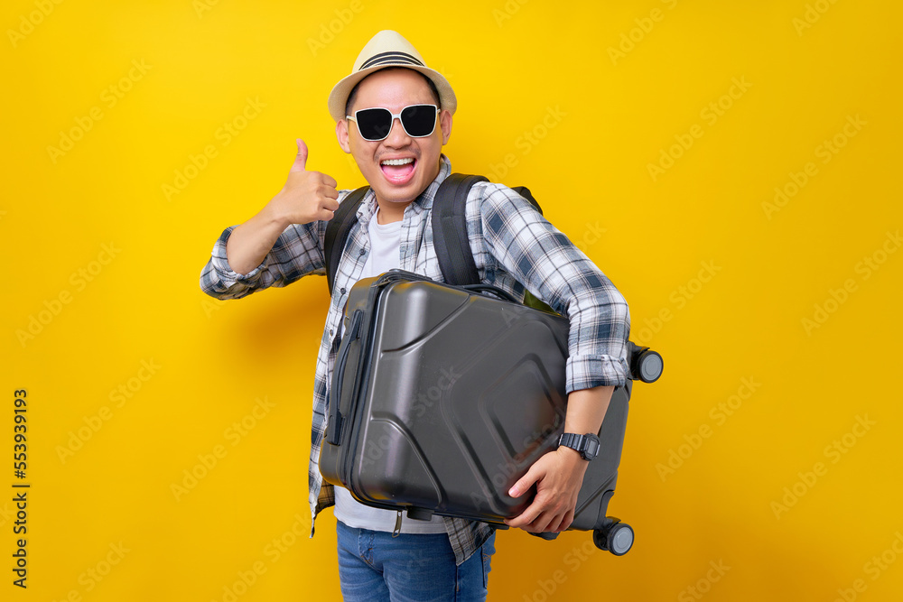 lets go. excited traveler tourist Asian man in casual clothes with hat glasses holding suitcase, showing thumb up on yellow background. Passenger traveling on weekends. Air flight journey concept lets go. excited traveler tourist Asian man in casual clothes with hat glasses holding suitcase, showing thumb up on yellow background. Passenger traveling on weekends. Air flight journey concept