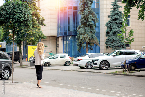 Banner with background urban modern landscape: business center, parking, cars, road, people. Woman with a phone view from the back. Summer. Сity ​​concept. Copy space on the right