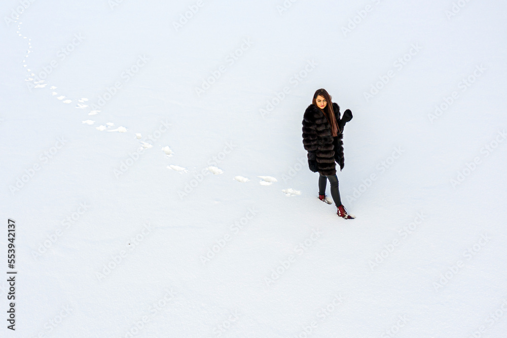 a cheerful girl in a warm fur coat walks across the open field leaving footpath in the snow