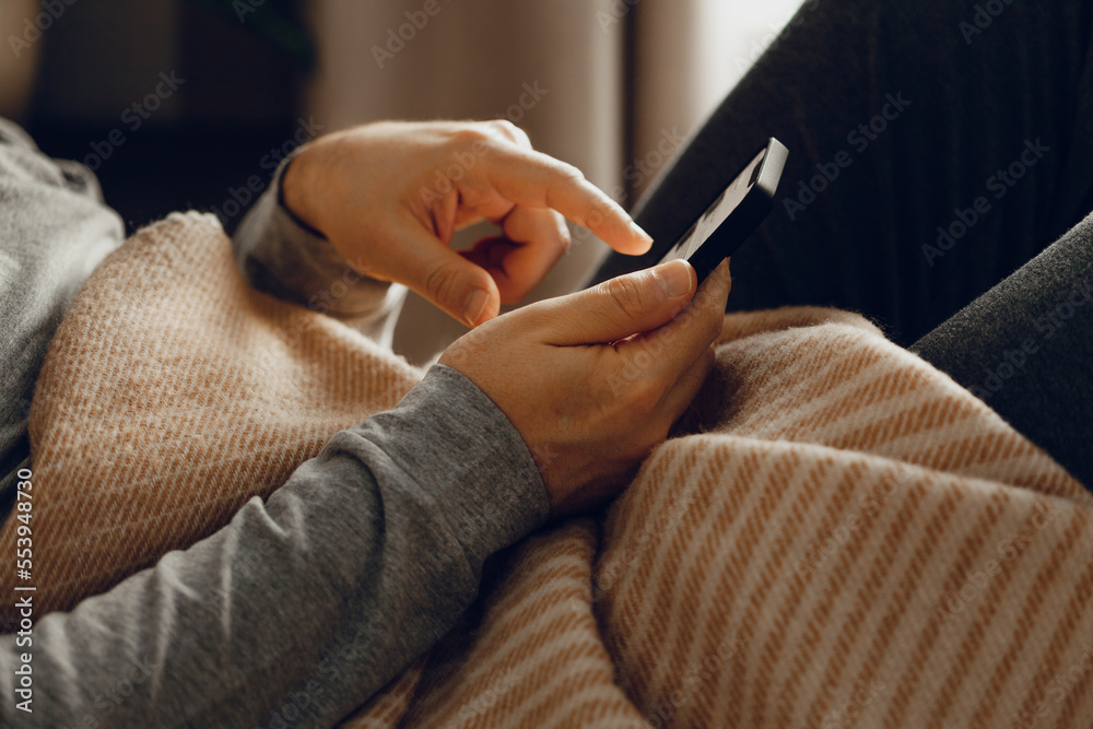 Fototapeta premium a caucasian man relaxing, using smart phone lying in bed under throw at home