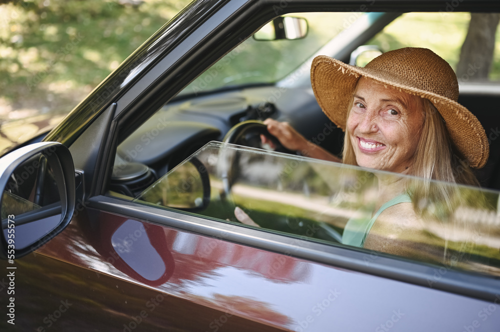 Fototapeta premium Happy senior woman driver in straw hat driving sitting in new car, smiling looking at camera enjoying journey. Driving courses and life insurance. Retired people activity and road trip concept 