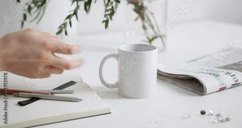 Person is placing  white colored coffee mug on a desk near the newspapers and note pad. photo