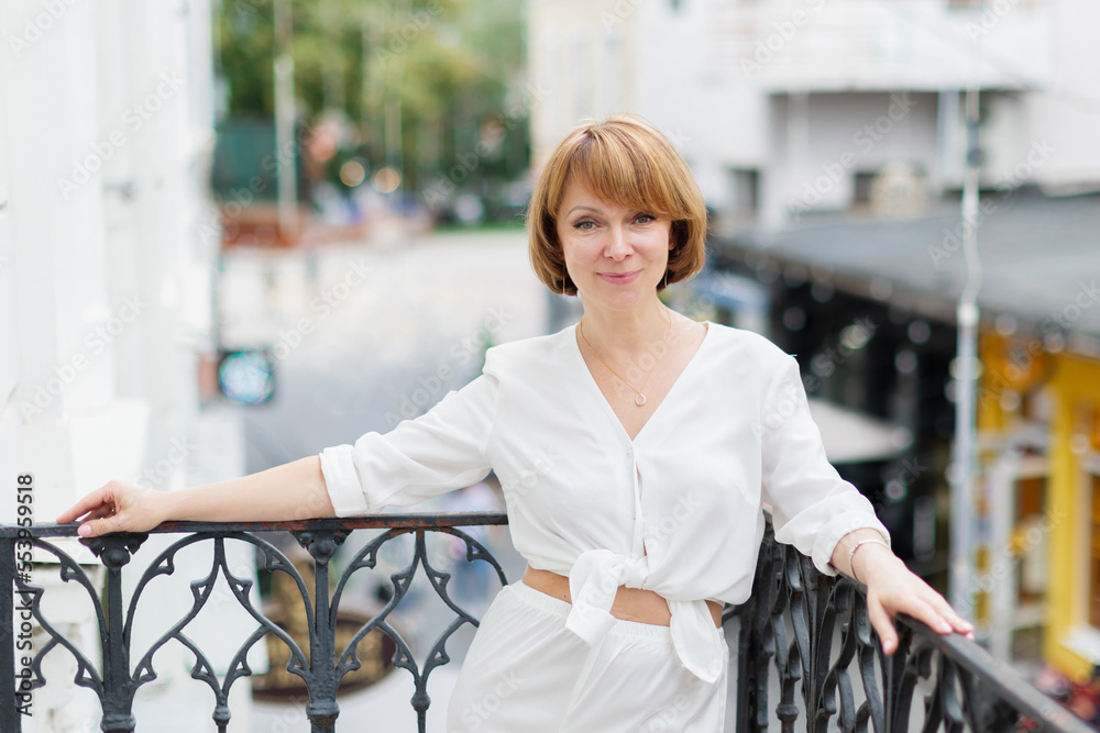 Adult woman in white suit is standing on balcony in town and looking at camera. Adult woman in white suit is standing on balcony in town and looking at camera.