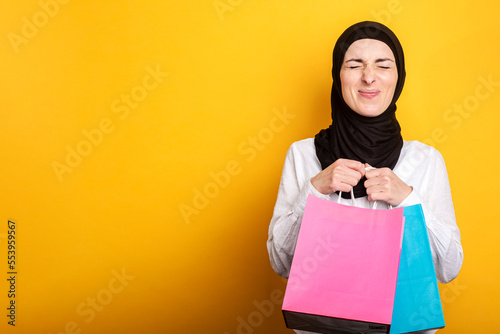 Young Muslim woman in hijab holds shopping bags, closed her eyes and rejoices on a yellow background. Banner