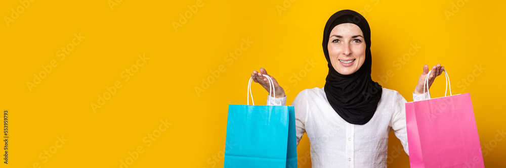Young muslim woman in hijab smiles and holds shopping bags on yellow background. Banner
