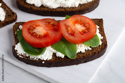 Slice of rye bread with cottage cheese and tomatoes on a wooden cutting board on a white background