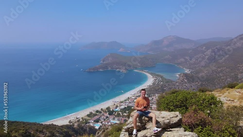 Panoramic view of man sit on the rock with remote control drone adjusting it, shooting himself aerial. Oludeniz beach photo