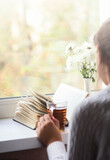 woman holding cup of tea and reading a book beside the window