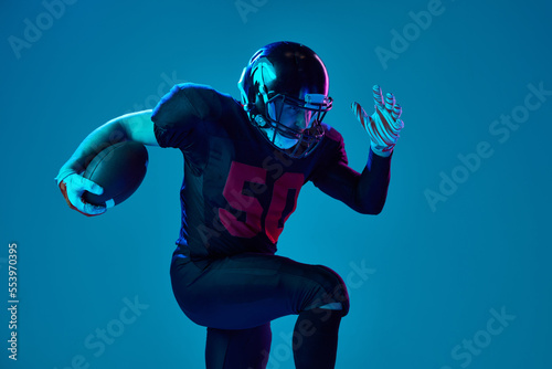 Sportive, strong man, american football player in sports team uniform and protective helmet isolated over blue background in neon light. Power, energy, achievements, skills