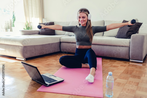 Shot of a sporty young woman using a laptop and doing lunges while dumbbells while exercising at home.