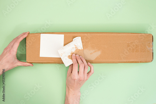 Hands peel off the label from a long paper parcel on a light green background. Product. Scam. Seal. Send. Shipment. Shipping. Stamp. Stealing. Sticker. Tag. Theft. Transportation. Trick photo