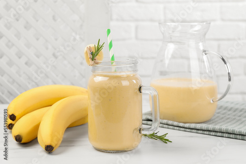 Mason jar and jug of tasty banana smoothie with fresh fruits on white marble table