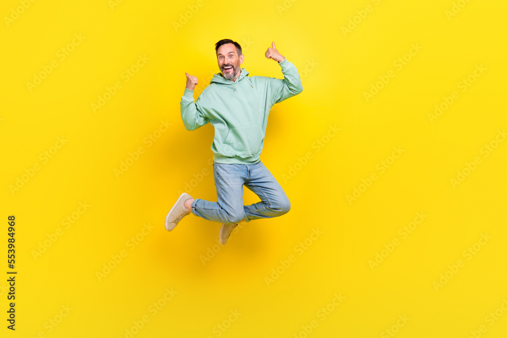 Full length photo of confident cool man wear green sweatshirt showing thumbs up jumping high isolated yellow color background