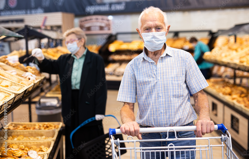 mature european man wearing mask and gloves with covid protection chooses buns and bread in supermarket bakery