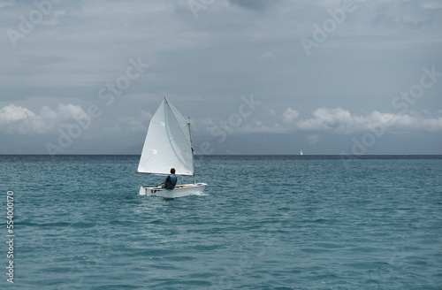Lonely sailor on training sailing pram optimist education boat in the sea in Greece, water background and cloudy sky © Ela
