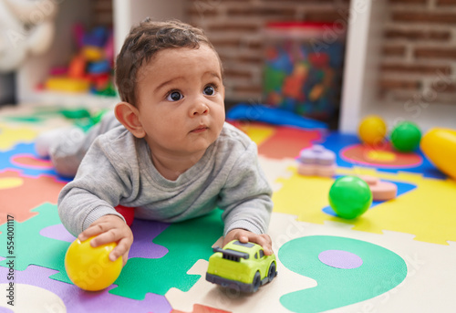 Adorable hispanic baby playing with ball and car lying on floor at kindergarten