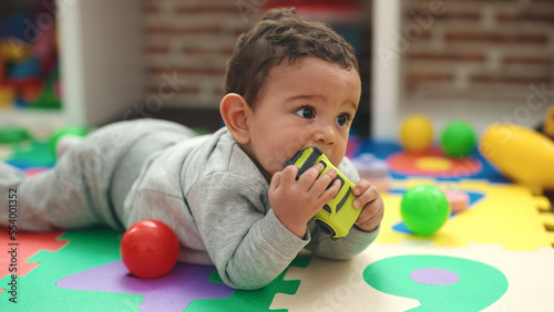 Adorable hispanic baby sucking car toy lying on floor at kindergarten